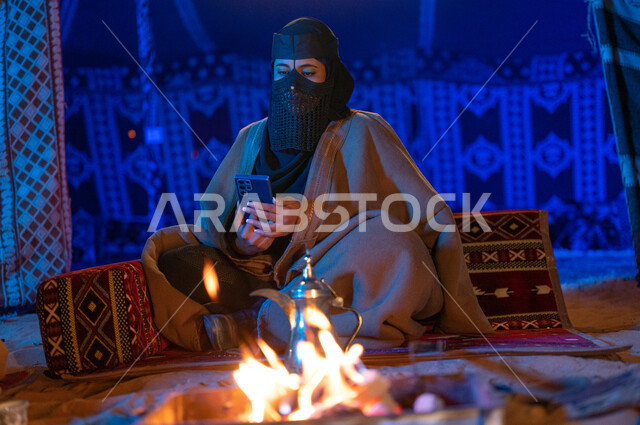 إشعال موقد النار أثناء التخييم في الصحراء السعودية، كشتة بر في الأجواء الشتوية الهادئة، امرأة عربية خليجية سعودية ترتدي البرقع والفروة تقوم باستخدام الهاتف الجوال داخل خيمة شعبية تراثية
