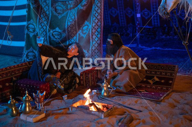 إشعال موقد النار أثناء التخييم في الصحراء، التقاط صور السيلفي التذكارية، عادات وتقاليد سعودية أصيلة في تقديم القهوة العربية، حسن الكرم والضيافة، كشتة بر في الأجواء الشتوية الهادئة، سيدتان عربيتان خليجيتان سعوديتان ترتديان الفروة تجلسان داخل خيمة شعبية تراثية