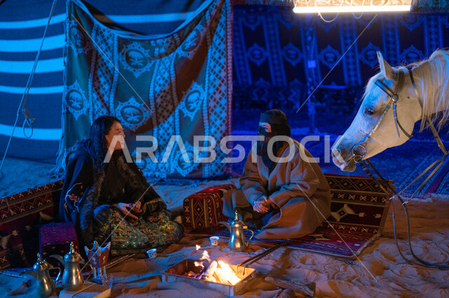 عادات وتقاليد سعودية أصيلة في تقديم القهوة العربية، مفهوم حسن الكرم والضيافة، إشعال موقد النار أثناء التخييم في الصحراء، كشتة بر في الأجواء الشتوية الهادئة، سيدتان عربيتان خليجيتان سعوديتان ترتديان الفروة تجلسان داخل خيمة شعبية تراثية
