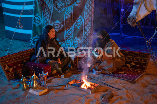 مفهوم حسن الكرم والضيافة، عادات وتقاليد سعودية أصيلة في تقديم القهوة العربية، إشعال موقد النار أثناء التخييم في الصحراء، كشتة بر في الأجواء الشتوية الهادئة، سيدتان عربيتان خليجيتان سعوديتان ترتديان الفروة تجلسان داخل خيمة شعبية تراثية
