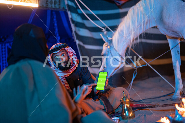 تربية الخيول الأصيلة، رجل خليجي سعودي يرتدي الشماغ يقوم باستخدام الهاتف الجوال بشاشة كروما خضراء فارغة، جلسة تخييم في الأجواء الشتوية في الصحراء السعودية، زوجان عربيان خليجيان سعوديان يرتديان الفروة يجلسان داخل خيمة شعبية تراثية ويستمتعان بشرب القهوة العربية