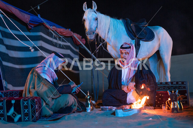رجل خليجي يقوم باستخدام الهاتف المحمول، تربية الخيول العربية الأصيلة، حسن الكرم والضيافة، إشعال موقد النار أثناء التخييم في الصحراء، كشتة بر شبابية في الأجواء الشتوية الهادئة، صديقان عربيان خليجيان سعوديان يرتديان الفروة والشماغ السعودي يجلسان داخل خيمة شعبية