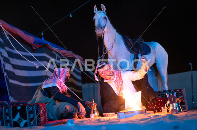 إشعال موقد النار أثناء التخييم في الصحراء، التقاط صور السيلفي التذكارية، مفهوم حسن الكرم والضيافة، كشتة بر في الأجواء الشتوية الهادئة، صديقان عربيان خليجيان سعوديان يرتديان الفروة والشماغ السعودي يجلسان داخل خيمة شعبية تراثية