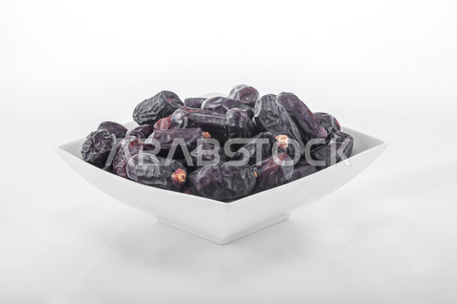 ضيافة رمضانية شهية، الأنواع المختلفة من التمور الصحية اللذيذة، غذاء صحي ومفيد، وجبة غذائية ذات أهمية عاليه، منتجات محلية سعودية وطنية، صورة مقربة لطبق أبيض مليء بحبات التمر السوداء، خلفية بيضاء 