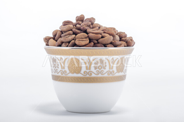 صنع المشروبات الساخنة  اللذيذة من حبوب البن، اعداد وتحضير القهوة الطازجة، جودة الصناعات والمحاصيل الغذائية الوطنية في المملكة العربية السعودية، صورة مقربة لفنجان ممتلئ ببذور وحبات البن المحمصة باللون البني، خلفية بيضاء.