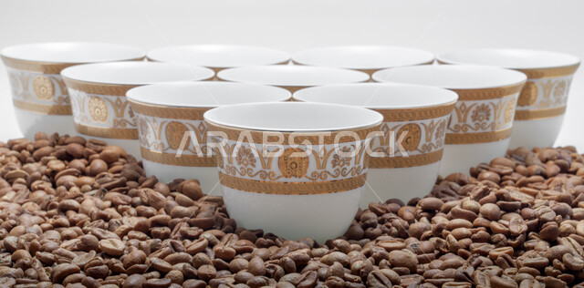 تحضير وإعداد القهوة الطازجة، جودة الصناعات والمحاصيل الغذائية الوطنية في المملكة العربية السعودية، صنع المشروبات الساخنة اللذيذة من حبوب البن، صورة مقربة لبذور وحبات البن المحمصة باللون البني المتناثرة حول الفناجين، خلفية بيضاء 