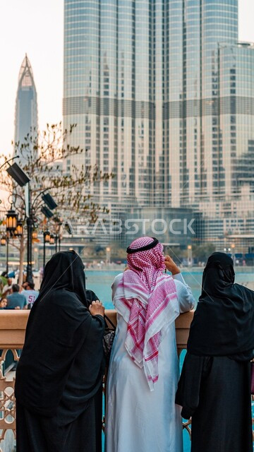 التقدم والنمو العمراني للأبراج وناطحات السحاب, صورة مقربة من الخلف لرجل عربي سعودي خليجي يرتدي الشماغ والثوب التقليدي مغ إمرأتان, إطلالة متميزة على البحيرات المائية, الاستمتاع والتأمل بالمناظر الطبيعية, السياحة واكتشاف المعالم المشهورة .
