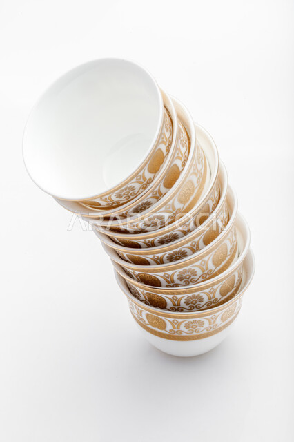 أكواب زجاجية صغيرة لتقديم المشروبات الساخنة، اكواب بيضاء مزخرفة باللون الذهبي ، صورة مقربة لمجموعة من الفناجين الصغيرة المرتبة فوق بعضها، فنجان ضيافة صغير، خلفية بيضاء