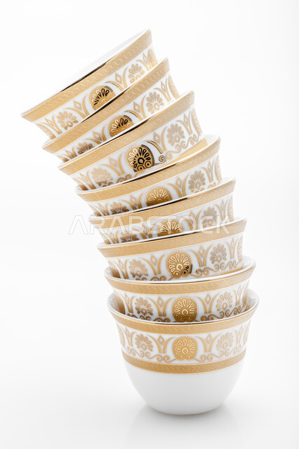 أكواب زجاجية صغيرة لتقديم المشروبات الساخنة، اكواب بيضاء مزخرفة باللون الذهبي ، صورة مقربة لمجموعة من الفناجين الصغيرة المرتبة فوق بعضها، فنجان ضيافة صغير، خلفية بيضاء