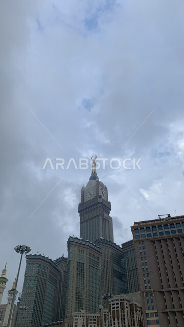 الفن الهندسي المعماري لناطحات السحاب، معالم دينية إسلامية مقدسة، صورة من الاسفل لمبنى برج الساعة الملكي في مكة المكرمة، العبادة والتقرب إلى الله، حجاج بيت الله الحرام في الحرم المكي الشريف في المملكة العربية السعودية، منظر السماء الملبدة بالغيوم
