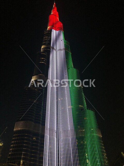 صورة مقربة لبرج خلفية المضاء بعلم دولة الامارات في وقت الليل، الاحتفال باليوم الوطني الاماراتي، التقدم والتطور المعماري في البلاد، معالم إمارة دبي السياحية