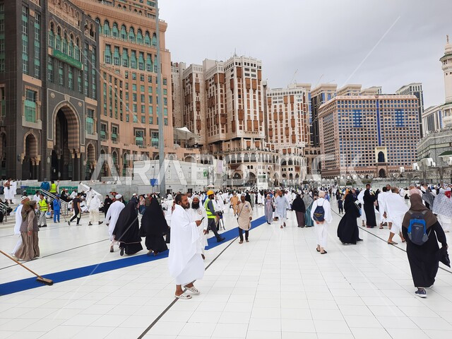 الحجاج والمعتمرين في الساحة الخارجية للحرم المكي في مكة المكرمه، التجمع الاسلامي من كل انحاء العالم لأداء العبادات والتقرب الى الله, الفنادق والأبراج الفاخرة المحيطة بالمسجد, أماكن ومعالم دينية اسلامية مقدسة, خلفية السماء الملبدة بالغيوم