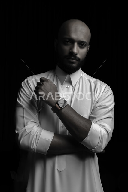 مفهوم الأناقة والرجولة والاهتمام بالمظهر الخارجي، بورتريه مقرب بالأسود والأبيض لشاب عربي خليجي سعودي يرتدي الثوب التقليدي وساعة اليد ينظر الى الكاميرا، تعابير وايماءات الجدية و الثقة بالنفس، خلفية سوداء