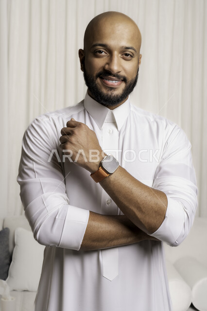 مفهوم الرجولة و الأناقة والاهتمام بالمظهر الخارجي، بورتريه مقرب لشاب عربي خليجي سعودي مبتسم يرتدي الثوب التقليدي وساعة اليد ينظر الى الكاميرا، تعابير وايماءات السرور و الثقة بالنفس، خلفية بيضاء