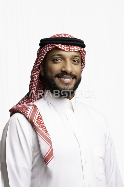 مفهوم الاناقة والرجولة، التقاط صور شخصية للوثائق الرسمية، الاعتناء والاهتمام بالمظهر الخارجي، بورتريه مقرب لرجل عربي خليجي سعودي مبتسم يرتدي الشماغ والثوب التقليدي يقف بأيدي متقاطعة ينظر الى الكاميرا بإيماءات السرور والثقة بالنفس، خلفية بيضاء .