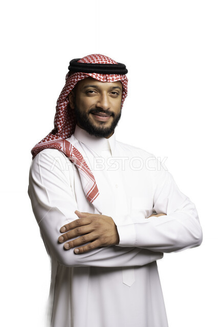 العناية والاهتمام بالمظهر الخارجي، التقاط صور شخصية للوثائق الرسمية، مفهوم الاناقة والرجولة، بورتريه مقرب لرجل عربي خليجي سعودي مبتسم يرتدي الشماغ والثوب التقليدي يقف بأيدي متقاطعة ينظر الى الكاميرا بإيماءات السرور والثقة بالنفس، خلفية بيضاء .