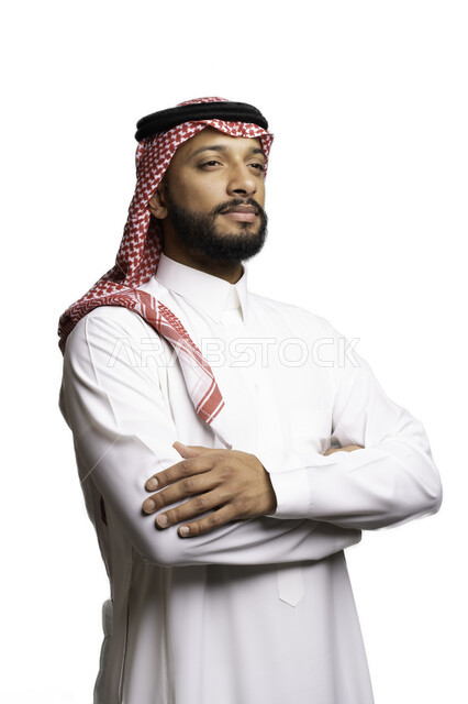 التركيز والتخطيط لحل مشكلة ما، مفهوم التردد والحيرة، الأناقة والاهتمام بالمظهر الخارجي، بورتريه لشاب عربي خليجي سعودي يرتدي الشماغ والثوب التقليدي يقف بحركات ايدي متقاطعة ينظر الى شيء ما بإيماءات التفكير لاتخاذ القرارات، خلفية بيضاء .