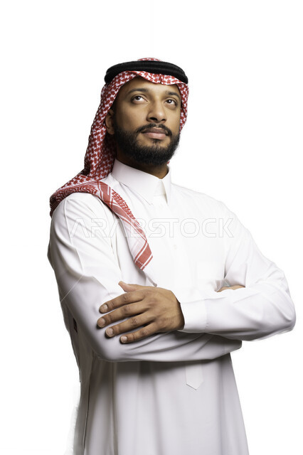 التفكير والتخطيط لحل مشكلة ما، مفهوم التردد والحيرة، الأناقة والاهتمام بالمظهر الخارجي، بورتريه لشاب عربي خليجي سعودي يرتدي الشماغ والثوب التقليدي يقف بحركات ايدي متقاطعة ينظر الى شيء ما بإيماءات التركيز لاتخاذ القرارات، خلفية بيضاء .