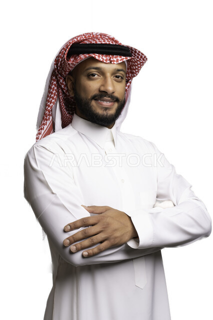 مفهوم الوسامة والرجولة، الأناقة والاهتمام بالمظهر الخارجي، التقاط صور شخصية للوثائق الرسمية، بورتريه مقرب لرجل عربي خليجي سعودي مبتسم يرتدي الشماغ والثوب التقليدي يقف بأيدي متقاطعة ينظر الى الكاميرا بتعابير السرور والثقة بالنفس، خلفية بيضاء .
