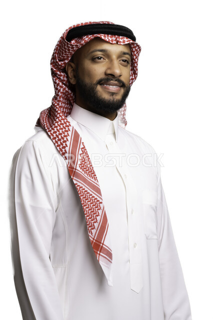 مفهوم التأمل والتركيز، إيماءات السرور والفرح، مفهوم الاناقة والرجولة، بورتريه مقرب لشاب عربي خليجي سعودي مبتسم يرتدي الشماغ والثوب التقليدي يقف باستقامة ينظر الى شيء ما، الاعتناء والاهتمام بالمظهر الخارجي، خلفية بيضاء .