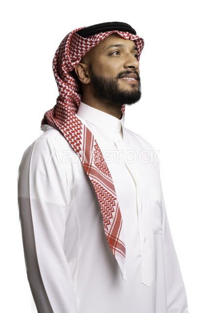 إيماءات السرور والفرح، مفهوم الاناقة والرجولة، بورتريه مقرب لشاب عربي خليجي سعودي مبتسم يرتدي الشماغ والثوب التقليدي يقف باستقامة ينظر الى شيء ما، الاعتناء والاهتمام بالمظهر الخارجي، خلفية بيضاء .