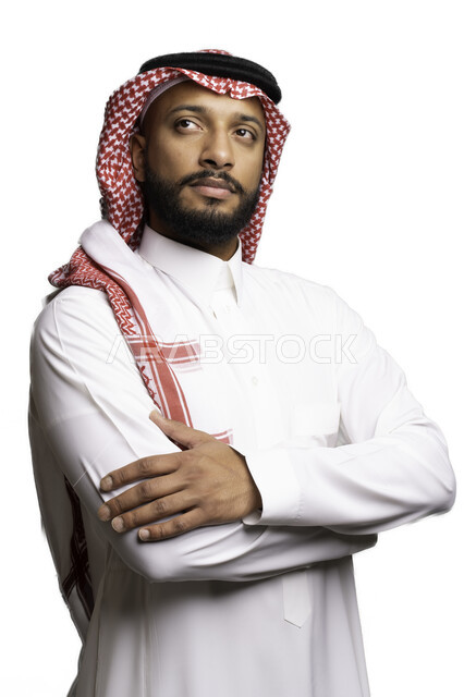 التمعن والتركيز لاتخاذ القرارات، مفهوم الحيرة والتردد، الأناقة والاهتمام بالمظهر الخارجي، بورتريه لشاب عربي خليجي سعودي يرتدي الشماغ والثوب التقليدي يقف بحركات ايدي متقاطعة ينظر الى شيء ما بإيماءات التفكير والتخطيط لحل مشكلة ما، خلفية بيضاء .