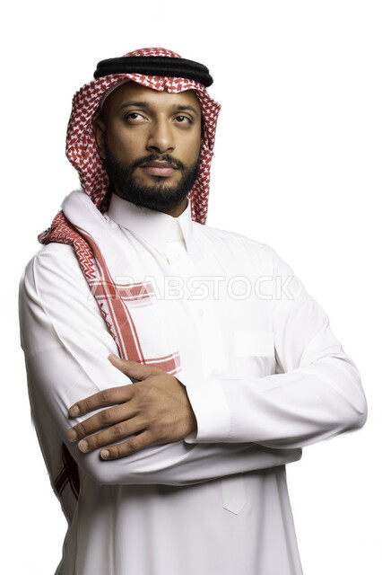 Focus and contemplation to make decisions, the concept of confusion and hesitation, elegance and attention to external appearance, a portrait of a Saudi Gulf Arab young man wearing a shemagh and traditional dress, standing with crossed hand movements, looking at something with gestures of thinking and planning to solve a problem, a white background