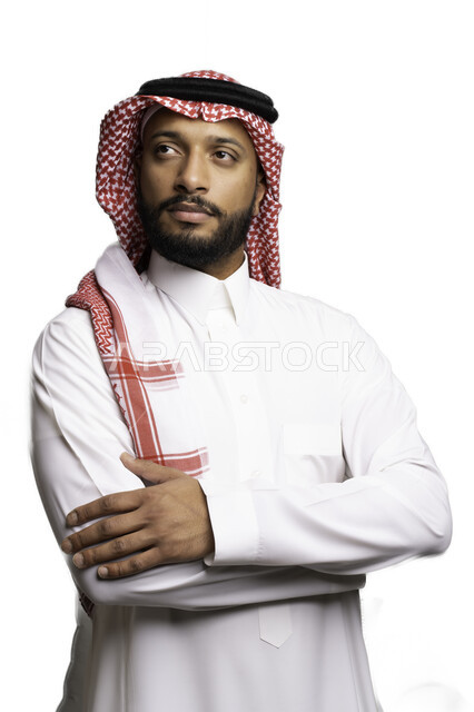 مفهوم الحيرة والتردد، التفكير والتخطيط لحل مشكلة ما، الأناقة والاهتمام بالمظهر الخارجي، بورتريه لشاب عربي خليجي سعودي يرتدي الشماغ والثوب التقليدي يقف بحركات ايدي متقاطعة ينظر الى شيء ما بإيماءات التركيز لاتخاذ القرارات، خلفية بيضاء .
