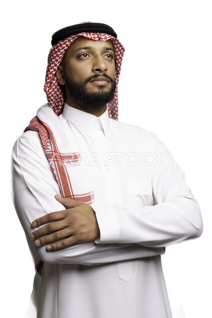مفهوم التردد والحيرة، التخطيط والتفكير لحل مشكلة ما، الأناقة والاهتمام بالمظهر الخارجي، بورتريه لشاب عربي خليجي سعودي يرتدي الشماغ والثوب التقليدي يقف بحركات ايدي متقاطعة ينظر الى شيء ما بإيماءات التركيز لاتخاذ القرارات، خلفية بيضاء .