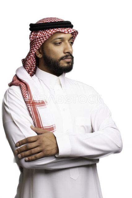 التفكير والتخطيط لحل مشكلة ما، مفهوم التردد والحيرة، الأناقة والاهتمام بالمظهر الخارجي، بورتريه لشاب عربي خليجي سعودي يرتدي الشماغ والثوب التقليدي يقف بحركات ايدي متقاطعة ينظر الى شيء ما بإيماءات التركيز لاتخاذ القرارات، خلفية بيضاء .