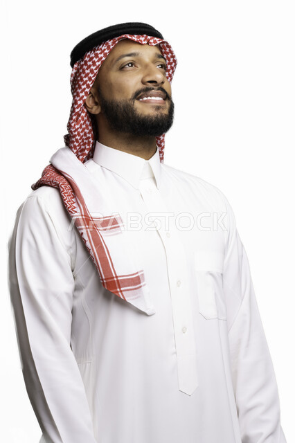 التأمل والنظر إلى شيء ما، إيماءات التفكير وشرود الذهن، مفهوم الاناقة والرجولة، بورتريه مقرب لرجل عربي خليجي سعودي مبتسم يرتدي الشماغ والثوب التقليدي يقف باستقامة ينظر إلى الأعلى، الاعتناء والاهتمام بالمظهر الخارجي، خلفية بيضاء .