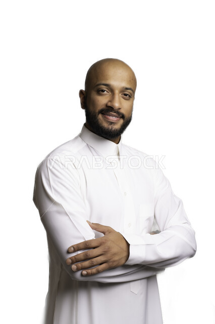 الاعتناء والاهتمام بالمظهر الخارجي، مفهوم الاناقة والرجولة، التقاط صور شخصية للوثائق الرسمية، بورتريه مقرب لشاب عربي خليجي سعودي أصلع مبتسم يرتدي الثوب التقليدي يقف بأيدي متقاطعة ينظر الى الكاميرا بتعابير السرور والثقة بالنفس، خلفية بيضاء .