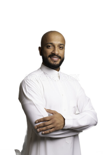 Taking care and attention to external appearance, the concept of elegance and masculinity, taking personal photos for official documents, a close-up portrait of a smiling, bald Saudi Gulf Arab young man wearing a traditional dress, standing with crossed hands looking at the camera with expressions of pleasure and self-confidence, white background