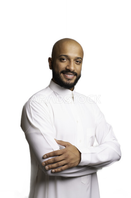 التقاط صور شخصية للوثائق الرسمية، الاعتناء والاهتمام بالمظهر الخارجي، مفهوم الاناقة والرجولة، بورتريه مقرب لرجل عربي خليجي سعودي أصلع مبتسم يرتدي الثوب التقليدي يقف بأيدي متقاطعة ينظر الى الكاميرا بإيماءات السرور والثقة بالنفس، خلفية بيضاء .