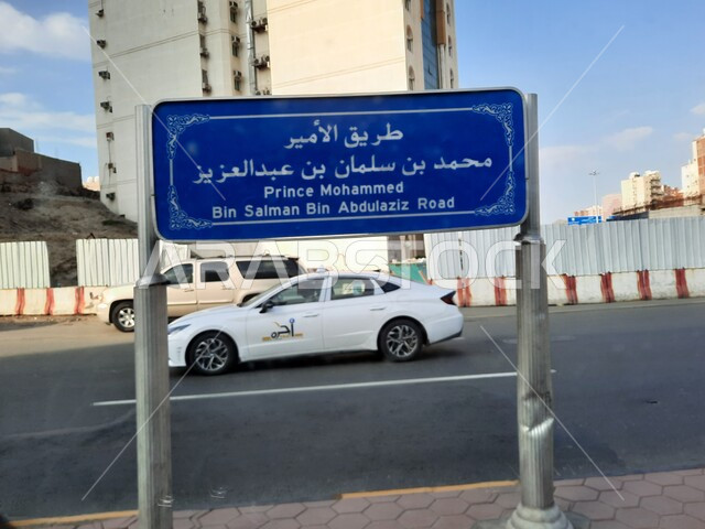 An explanatory sign for Prince Mohammed bin Salman bin Abdulaziz Road, sacred Islamic religious landmarks and places, interest in construction and urban development in Mecca, car traffic on paved roads and streets in the Kingdom of Saudi Arabia, luxury hotels and towers surrounding the Grand Mosque