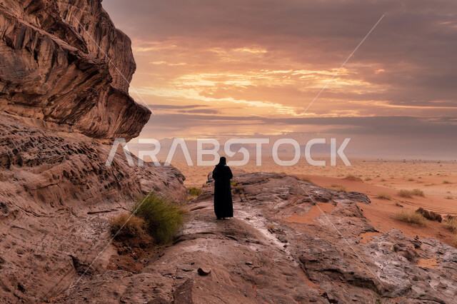 التكوينات الصخرية في منطقة نيوم في المملكة، الاستمتاع بالمناظر الطبيعية في وقت غروب الشمس، سلسة جبلية في مدينة تبوك الصحراوية، معالم سياحية مشهورة, صورة من الخلف لامرأة عربية خليجية سعودية محجبة تتأمل التضاريس والنحت الصخري في المناطق المرتفعة