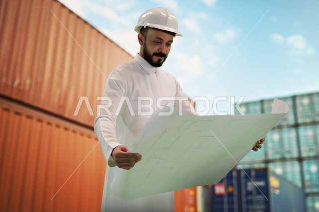 Working in the engineering sector, the concept of engineering and construction, supervising the implementation of architectural plans and projects in Saudi Arabia, a smiling Saudi Gulf Arab engineer with a protection helmet holding a building plan in his hand standing at his workplace