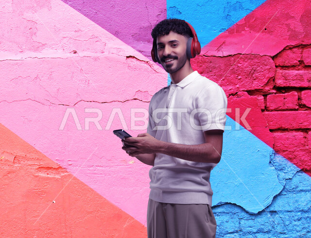 Listening to songs using a headphone, using audio programs on a mobile phone, a portrait of a smiling Saudi Gulf Arab young man wearing a casual outfit, wearing headphones, holding a mobile phone in his hand, looking at the camera with happy gestures, a background of a colored stone wall.