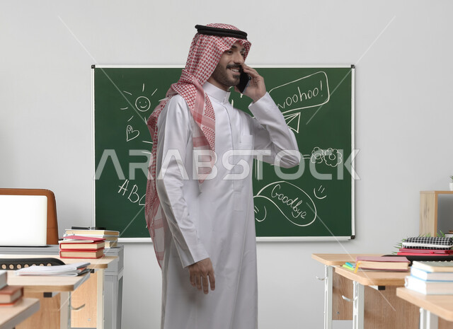 معلم عربي سعودي خليجي يرتدي الشماغ والثوب التقليدي يقوم بإجراء مكالمة هاتفية باستخدام الهاتف المحمول بإيماءات السرور، نظام التعليم في المملكة العربية السعودية, انتظار موعد الدرس، خلفية السبورة في الفصل الدراسي