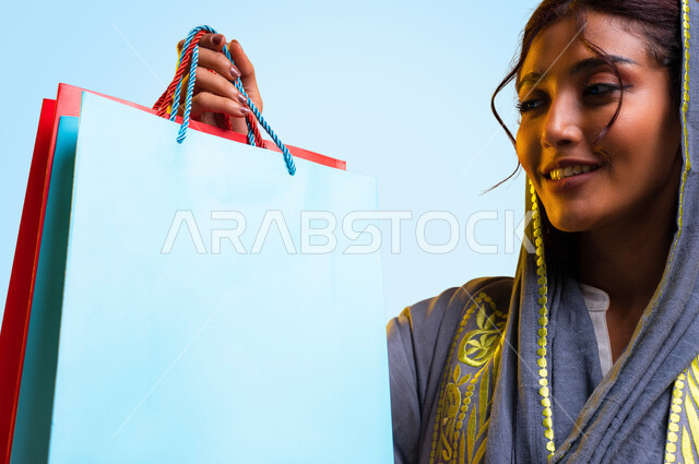 السعادة والتأثير الإيجابي على السيدات في زيارة المولات التجارية في السعودية، الاستفادة من العروض الموسمية والتخفيضات, التبضع وشراء المستلزمات، بورتريه مقرب لامرأة عربيه سعودية خليجية ترتدي العباءه تحمل بيدها أكياس التسوق الملونة، خلفية زرقاء