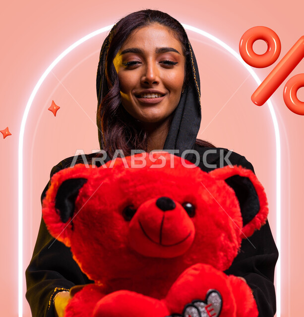 ايماءات الفرح والسعادة بالحصول على هدية بمناسبة الأعياد والمناسبات السعيدة، رمز الحب والرومانسية، بورتريه مقرب لسيدة عربية خليجية سعودية مبتسمة ترتدي عباءة سوداء تحمل بيدها دبدوب أحمر, خلفية ملونة