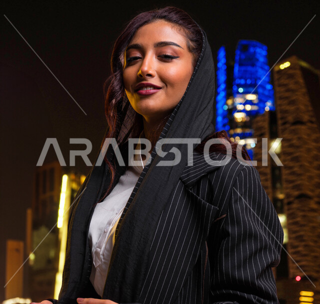 مفهوم الأنوثة والنعومة، ارتداء أحدث تصاميم الملابس النسائية السعودية، الاهتمام بالجمال والأناقة، صورة مقربة لشابة عربية سعودية خليجية مبتسمة ترتدي جاكيت أسود مخطط تنظر إلى الكاميرا بإيماءات السرور، متابعة أخبار وصيحات الموضة، خلفية الأبراج المضاءة ليلا