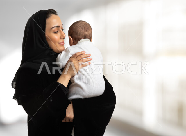 مفهوم حنان الأمهات، تحمل المسؤولية والعناية بالأطفال، قضاء وقت ممتع مع البيبي، صورة مقربة لأم عربية خليجية سعودية محجبة مبتسمة تحمل طفلها الرضيع تحضنه بعطف وحب، مشاعر الأمومة والطفولة، خلفية بيضاء