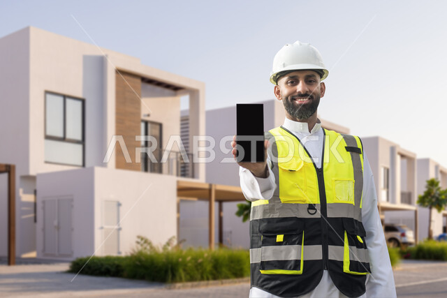 مهندس عربي خليجي سعودي مبتسم يرتدي خوذة وسترة الحماية الخاصة بالعمل يعرض شاشة سوداء فارغة عبر الهاتف المحمول، الإشراف على انشاء المباني في السعودية، استخدام التقنية والتكنولوجيا في المشاريع المستقبلية، خلفية شاليهات وشقق سكنية