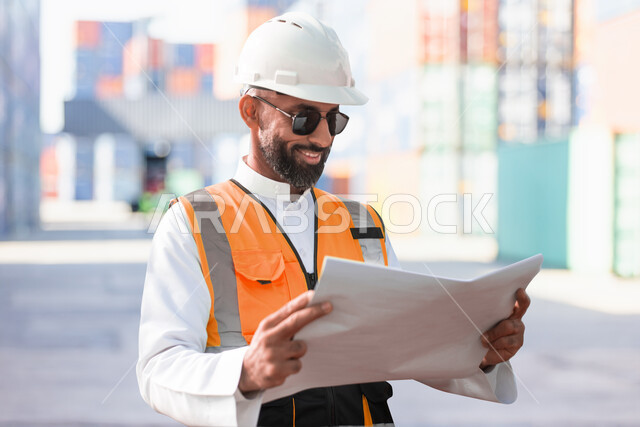 متابعة المشروع وتدقيق المخططات، مهندس معماري عربي خليجي سعودي مبتسم يرتدي النظارات الشمسية وخوذة وسترة الوقاية الخاصة بالعمل يحمل بيده مخطط البناء، الاشراف والرقابة على الانشاءات الهندسية، التحقق من تطبيق المشاريع والخطط الهندسيه المعمارية في البناء بشكل صحيح