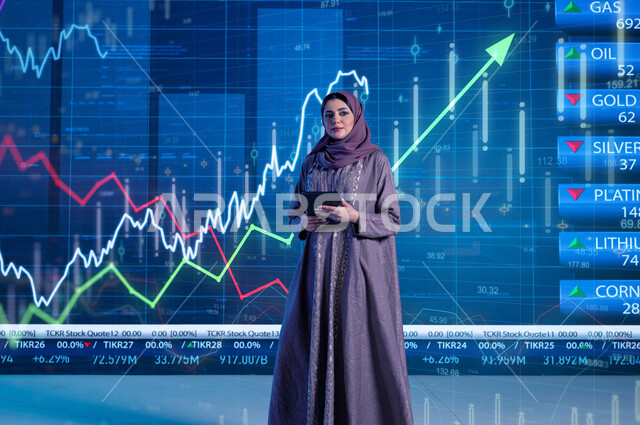 توضيح التغيرات الحاصلة في أسعار السلع النفطية والثروات، مذيعة وإعلامية عربية خليجية سعودية ترتدي الحجاب والعباءة تقوم بتقديم النشرات الإخبارية الإقتصادية، قراءة الموجز الأخباري بإستخدام جهاز التابلت، خلفية شاشة إلكترونية حديثة لصعود وهبوط مؤشر الأسهم والبورصة