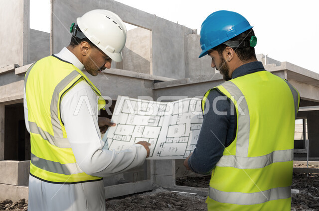 متابعة المشروع وتدقيق المخططات، صورة من الخلف لمهندسين عربيين خليجيين سعوديين يرتديان خوذة وسترة الوقاية الخاصة بالعمل يتناقشان حول مخطط البناء، الاشراف والرقابة على الانشاءات الهندسية، التحقق من التطبيق الصحيح للمشاريع المعمارية، خلفية موقع العمل