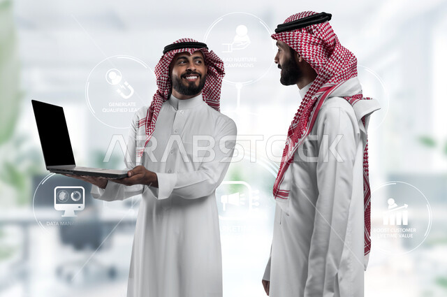 متابعة مشاريع الأعمال على الحاسب المحمول، صورة مقربة لرجلي أعمال سعوديين خليجيين عربيين يرتديان الشماغ والثوب التقليدي يقومان بإنجاز مهام العمل عبر جهاز الحاسوب، التعاون بين الموظفين لتحقيق أهداف الشركة، خلفية أيقونات مكتبية بتقنية الهولوجرام