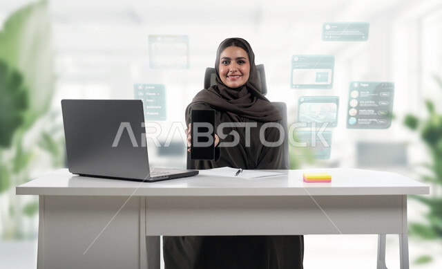 Displaying commercial applications via mobile phone, a veiled Saudi Arabian Gulf woman wearing an abaya sitting behind the table and holding a mobile phone with a blank black screen in her hand, integrating technology and technology into practical life, office background with task and business icons using hologram technology