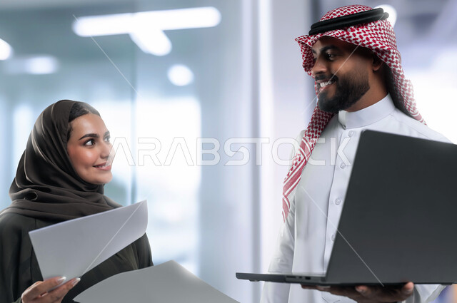 تطوير الاستراتيجيات وتبادل الأفكار والخبرات لتحقيق أهداف العمل، التعاون وروح الفريق الواحد، بورتريه مقرب لزميلين عربيين سعوديين خليجيين يقومان بمناقشة  وتنظيم الاعمال بإستخدام جهاز الحاسوب المحمول، دمج العمل مع التقنية والتكنولوجيا، خلفية المكتب ضمن مقر الشركة